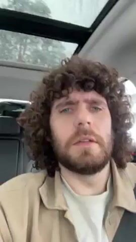 The video features a man with curly brown hair and a beard sitting in a car. He is wearing a white shirt and a beige jacket. The car has a sunroof. The man is talking and gesturing. The background is blurry, but it appears to be a snowy day.