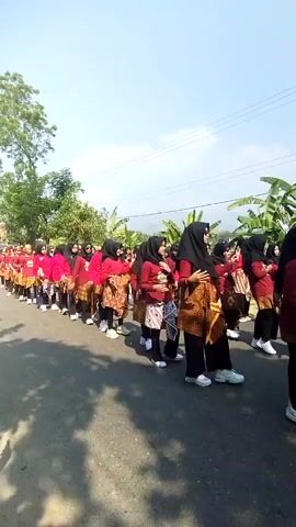 The video shows a group of people, predominantly women, walking in a single file line on a paved road. They are dressed in matching red tops and patterned skirts, with some wearing headscarves. The individuals are walking forward, appearing to be participating in some kind of procession or parade. The environment appears to be outdoors, with trees, foliage, and a bright, sunny sky visible. The road is asphalt and seems to be a rural or semi-rural area, as evidenced by the natural surroundings. The perspective of the video is from someone walking behind and slightly to the side of the group.