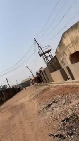The video shows a street view in what appears to be a developing or rural area. The road is unpaved and dirt-covered. Buildings on either side of the road are simple, some appearing worn. There are power lines strung across the sky, supported by wooden poles. Birds can be seen flying in the sky and perched on the power lines. The sky is a clear, pale blue.