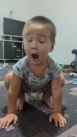 A young child, wearing a light-colored t-shirt with a print and a diaper, is shown in various positions on the floor and on a sofa. The child is first seen in a squatting position on a carpeted floor, then crawling on all fours. The child then climbs onto a sofa, where toys are visible. The environment appears to be an indoor living space with a television set in the background and other furnishings. The child's movements are observed as they explore their surroundings.
