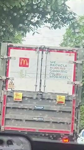 From the perspective of a driver, the video shows the back of a McDonald’s truck on a rainy day. The truck’s door has “We recycle all our used cooking oil into biodiesel” printed on it. There are trees flanking either side of the road and other vehicles are visible in the distance.
