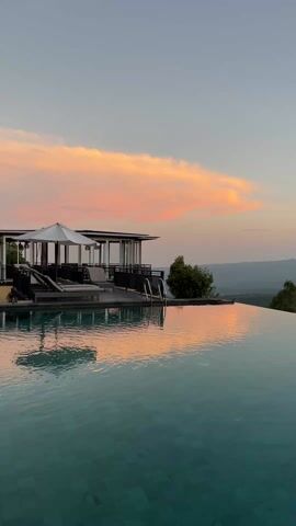 The video shows an infinity pool with a scenic mountain view during sunset. The sky is painted with soft hues of pink and orange, with a cloud adding to the drama. The sun’s glow is reflected on the water, adding a tranquil touch to the scene. On the edge of the pool is a pavilion-like structure with lounge chairs. The scene evokes a sense of serenity and luxury.
