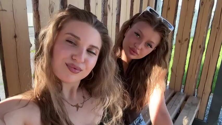 Two young women with light brown hair, wearing sunglasses, are posing for a selfie. They are sitting close together on a wooden bench in front of a slatted wooden structure. Music is playing in the background. The woman on the left has a tattoo on her chest and is wearing a necklace. She winks and sticks out her tongue. The woman on the right makes a kissy face and a peace sign. The background shows green grass and possibly a building.