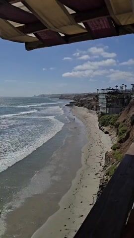 An aerial shot shows a scenic beach with waves crashing on the shore. A cliffside path overlooks the beach, with a building perched on top. Two people are walking along the beach. The sky is blue with scattered clouds.