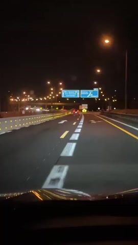 The video shows a view from inside a vehicle moving on a highway at night. The road is well-lit by streetlights and the car's headlights. Lane markings are visible, and a sign indicates directions to Wien (Vienna), Budapest, Graz, Eisenstadt, Brunn, and Campus 21. There are other vehicles on the road, but they are not close to the camera car. A temporary road barrier with reflective elements lines the left side of the road.