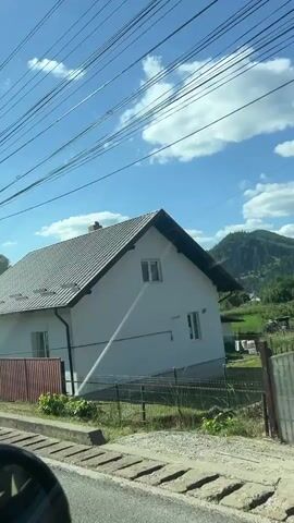 The video shows a view from inside a vehicle traveling on a road. The first scene is of a white house with a gray tiled roof. A stream of water is being sprayed from a yellow hose onto the side of the house. Next, the view shifts to a gray house with a dark gray tiled roof. Then, a white church with a tall steeple and a red door comes into view. Finally, the video shows a utility pole with wires and a cluster of trees. The sky is blue with scattered clouds.
