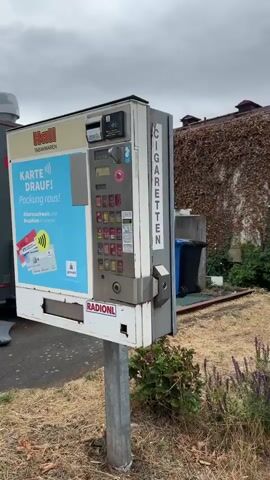 The video shows a close-up view of a cigarette vending machine. The machine is silver and gray with various cigarette brands and prices displayed in rows. There are buttons for selecting cigarettes, a coin slot, and a slot for banknotes. The machine is located outdoors, with a wall covered in ivy in the background and some dry grass and plants in the foreground. In one shot, a hand with manicured fingernails is seen holding a plastic bottle near the machine. The machine has a sign in German that says "ZIGARETTEN MARKE" and "Rauchen ist tödlich" (Smoking is deadly). There is also a warning about the graphic images on cigarette packs. The video appears to be shot in handheld style, with some slight camera shake.