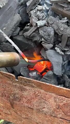 A close-up shot shows a piece of metal being heated over glowing coals in a grill. Sparks fly from the metal as it makes contact with the hot embers. The coals are black and gray, with bright orange and red spots where they are burning. Smoke rises from the grill. The grill itself appears to be made of rusted metal. A wire rack is visible above the coals. The background is blurred, suggesting an outdoor setting.