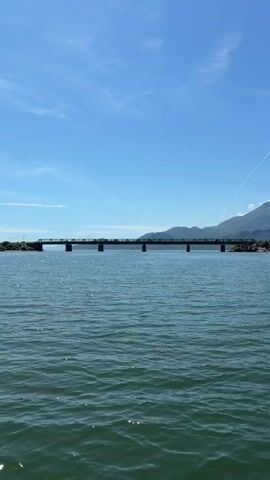 The video shows a wide shot of a body of water with a long, low-lying bridge spanning across it in the mid-ground. The water is a deep green-blue and has gentle ripples reflecting sunlight. In the background, beyond the bridge, are distant mountains under a clear, bright blue sky with a few wispy clouds. The camera is moving forward smoothly, suggesting it is on a boat or floating object, heading towards the bridge. The overall scene is bright and sunny.