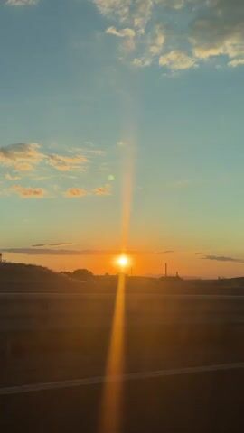 The video captures a vertical shot of a sunset as seen from a vehicle moving on a road. The sun is a bright orb on the horizon, casting a strong lens flare that extends upwards. The sky is a gradient of blue and orange, with scattered white clouds. In the foreground, a dark road surface with a white line is visible, separated from the background by a concrete barrier. Trees and distant structures line the horizon. The footage appears to be a continuous shot with slight camera shake, indicating movement.