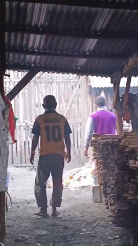 The video shows two men walking away from the camera inside a covered outdoor area. The first man is wearing a yellow shirt with the number 10 on the back and dark pants. The second man is wearing a purple jacket and light-colored shorts. There are stacks of wood to the right of the frame and a fire burning in the background. The ground is dirt and debris.