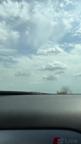 A first-person perspective video shows a drive on a two-lane road through a rural area. The sky is cloudy with patches of blue. Trees and shrubs line the road. A white car is visible in the distance. The dashboard of the vehicle is visible in the foreground.