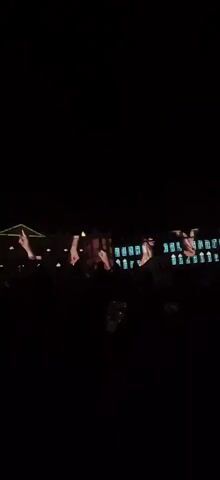 The video shows a nighttime outdoor scene with a crowd of people visible in silhouette in the foreground. Behind them, a building is illuminated with a projection of animated hands and abstract patterns, creating a light show. The hands appear to be making gestures, and the patterns change color and form throughout the clip. The overall impression is of a public event or performance taking place at night.