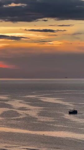 The video captures a serene sunset over the ocean in Lý Sơn Special Administrative Region, Vietnam. The sky is a dramatic canvas of deep oranges and yellows, tinged with soft purples and grays from scattered clouds. The sun's warm glow illuminates the horizon, casting a gentle light across the vast expanse of the sea. The water is calm, with subtle patterns created by gentle waves reflecting the colors of the sky.

A lone fishing boat, dark in silhouette, sails across the middle ground, its wake trailing behind it. In the distance, several other vessels are visible as small specks on the horizon, suggesting a moderately active shipping lane or fishing area. The overall mood is one of peaceful solitude and natural beauty. The scene is likely set in the late afternoon or early evening, as indicated by the characteristic colors of a sunset. There is no visible text in the video.