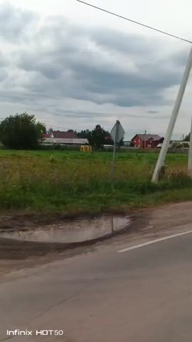 The video shows a view from a moving vehicle looking out at a rural landscape under a cloudy sky. A paved road with a white line runs along the bottom of the frame. To the left of the road, there is a dirt shoulder and a grassy field with scattered wildflowers and some yellow structures. In the background, there are several houses, trees, and utility poles. One house in the distance is red. The camera appears to be positioned to the right of a tall, light-colored utility pole. The time stamp at the bottom indicates it was recorded on an Infinix HOT 50.