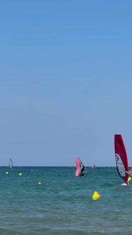 The video shows a bright, sunny day with a clear blue sky over a calm ocean. Several people are windsurfing. In the foreground, a person on a windsurf board with a large red sail is actively engaged in the sport. In the middle ground, another person is windsurfing with a pink and white sail. Further in the distance, there are other windsurfers and some buoys on the water. The water is a deep blue-green. The scene appears to be a popular spot for water sports.