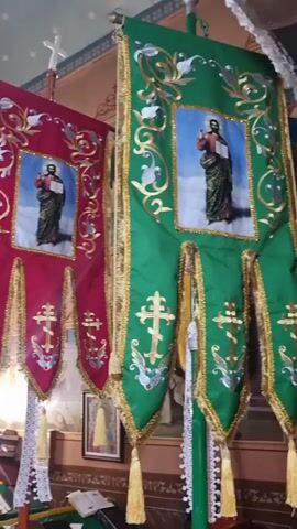 This media file captures a dimly lit interior of an Orthodox church. The primary subjects are two large, ornate banners hanging from a wooden pole. The banner on the left is a deep red, and the one on the right is green. Both banners feature golden embroidery and depict religious imagery. Each banner has a central square with a painting of Jesus Christ holding an open book, and below this, three hanging tassels with a cross design.

In the background, a gilded iconostasis, a common feature in Orthodox churches, is visible. It is adorned with numerous religious icons, including depictions of saints and Christ. A woman with reddish-brown hair is seen from behind, holding a smartphone, seemingly taking a photograph or recording video of the church's interior. A large, elaborate chandelier hangs from the ceiling, casting a warm light. The overall atmosphere appears solemn and reverent, characteristic of a place of worship. The setting is clearly an Orthodox church, and given the Ukrainian context, it's likely a traditional place of worship within the country. The time of day is not discernible from the lighting alone, but the indoor setting suggests it could be any time. No text is visibly legible within the frame.