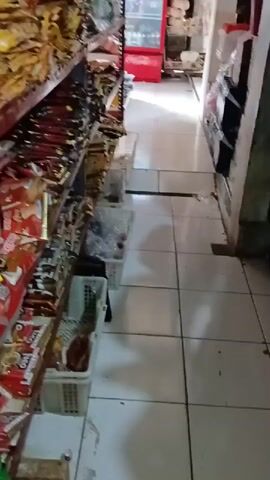 The video shows a narrow aisle within a store. Shelves filled with packaged goods, possibly snacks or groceries, line the left side of the aisle. The floor is tiled with white and gray squares, and some of the tiles appear worn or damaged. On the right side of the aisle, a wooden crate filled with brown eggs is visible. A metal bar or rod is propped against the crate. The overall impression is of a small, possibly local, retail establishment.