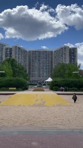 The video shows a playground with a large sand pit in front of a multi-story residential building on a partly cloudy day. The sand pit has a raised yellow section in the center. Two children are playing in the sand pit, running and walking around. People are sitting on benches near the sand pit. Trees and bushes surround the playground, and a paved brick path is visible in the foreground. A white tent-like structure is situated behind the yellow structure and in front of the buildings.