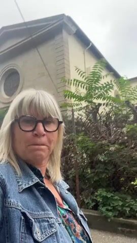 A woman with blonde hair and black-rimmed glasses is walking towards the camera in front of a building. She is wearing a denim jacket over a colorful patterned shirt. To her right is a fence and some foliage. Behind her to the left, a car is parked on the side of the road. The sky is overcast.