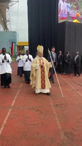 A religious procession is taking place outdoors on a reddish-brown track, likely a stadium, under an overcast sky. The central figure is a bishop in golden vestments, a mitre, and holding a crosier, walking towards the right. He is followed by other clergy in white and black, and white and magenta vestments, some with hands clasped in prayer. Several men in dark suits, possibly security or officials, accompany the procession.

In the background, a large screen displays images of what appears to be a religious ceremony with numerous people in white clerical attire. Behind the screen, black fabric partitions are set up. To the right, a red carpet leads towards a partially visible black Mercedes-Benz van. The individuals in the procession appear solemn as they move forward.