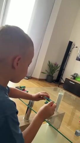A young boy, viewed from behind and slightly to the side, is sitting at a glass-topped coffee table, engaged in playing with colorful building blocks, possibly LEGOs. He is wearing a blue t-shirt. The boy has short, light-brown hair. His hands are actively manipulating the blocks on the table.

The scene is indoors, suggesting a home environment. The floor is made of light-colored wood. In the background, a tall, dark speaker is visible, along with a black cabinet. A potted plant with green leaves and pink flowers sits on top of the cabinet. A window with white curtains is to the left of the frame, letting in natural light, indicating it is daytime. The overall atmosphere appears to be one of focused play and concentration. No text is visible in the media.