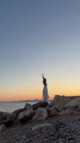 This is a vertical, eye-level shot of a lone woman standing on a rocky shore at sunset. She is wearing a long, flowing white dress and has her arms raised above her head, her hands clasped. Her back is to the camera, and her long, dark hair is cascading down her back.

The scene is set at dusk, with a pale crescent moon visible in the clear, gradient sky that transitions from a deep blue at the top to soft oranges and yellows near the horizon. The ocean is calm, with gentle waves lapping at the shore. In the distance, silhouetted hills are visible. The foreground is dominated by a jumble of large, weathered rocks, interspersed with smaller pebbles and dark sand.

The overall mood is one of serene contemplation and peacefulness, perhaps even a sense of awe at the natural beauty of the sunset. The woman's pose suggests a moment of reflection or expression of joy. The location is identifiable as Paleo Faliro, Greece, a coastal area known for its beaches and seaside promenades. There is no visible text in the media.