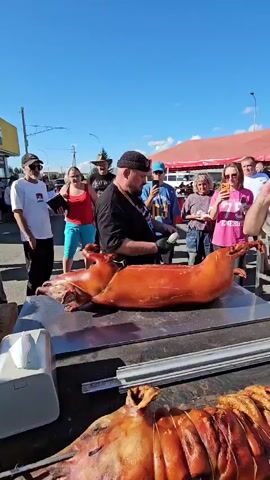 The video shows an outdoor event with a clear blue sky. In the foreground, a whole roasted pig is laid out on a metal table. Another roasted pig is partially visible at the bottom of the frame. Several people are gathered around the table, watching a man who is holding a knife and appears to be carving or preparing the pig. He is wearing a black apron over a black t-shirt and a black cap. He is also holding a small black device, possibly a microphone or a recording device, in his other hand. Some spectators are holding up their phones, suggesting they are recording or taking pictures. To the left of the main table, there is a white dispenser, possibly for napkins or wipes. In the background, there are tents, a building with a yellow sign, and other people milling about. The overall atmosphere appears to be a food event or a gathering. The man speaking is heard to say "It actually took us about an hour and a half to cook."