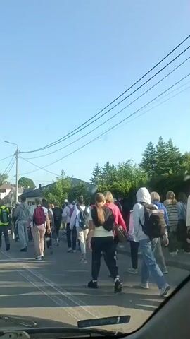 A group of people, appearing to be students and adults, are walking in a line on a paved road. Most of them are wearing backpacks and appear to be heading in the same direction. The environment is outdoors with a clear blue sky. Buildings and trees are visible in the background, suggesting a suburban or rural setting. The sun is shining, casting shadows on the road. The video is shot from a vehicle's perspective, looking forward.