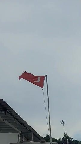 The video captures a Turkish flag flying in the wind against a cloudy sky. The flag is red with a white crescent moon and star. It is attached to a tall, silver flagpole. In the lower left corner of the frame, a section of a white building with a dark, sloped roof is visible. In the lower right corner, a lamppost and some green trees can be seen. The camera is positioned looking upwards.
