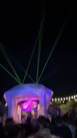 Here is a detailed description of the video:

**Scene:** The video captures a vibrant outdoor music event at night. The focal point is a stage structure with a unique geometric design.

**Stage Structure:** The stage appears to be a temporary, collapsible setup. It's illuminated with purple and green lighting, creating a party atmosphere. The front of the stage has a sign that reads "MONSTER MASH."

**Lighting and Special Effects:** Green laser beams shoot upward from the top of the stage, adding to the spectacle. There are also string lights in the background, further enhancing the festive ambiance.

**Crowd:** A dense crowd of people is gathered in front of the stage.

**Actions and Interactions:** People are standing, talking, and some are recording the scene with their phones. The music suggests dancing and enjoyment.

**Environment:** The event takes place outdoors under a clear, dark night sky.

**Overall Impression:** The video conveys a lively and energetic scene at a music event, possibly a festival or outdoor concert. The lighting, lasers, and crowd indicate a festive atmosphere.