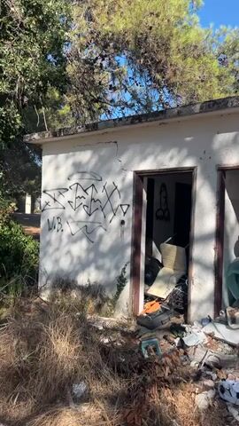 The video shows an exterior view of a small, dilapidated building with two open doorways. The building's exterior walls are white and covered in graffiti. The area in front of the building is overgrown with dry grass and littered with various debris, including trash, plastic bags, a soccer ball, and what appears to be discarded furniture and household items. The doorways lead into dark interiors that are also filled with clutter and debris, suggesting neglect and abandonment. Trees with green foliage are visible in the background against a clear blue sky.