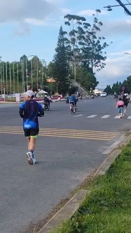 A man in athletic wear runs across a street with a pedestrian crossing. He is wearing a blue and black shirt, black shorts, a white hat, and white shoes. In the background, several motorcycles and bicycles are visible, as well as other pedestrians. The street is paved, and there is grass on the right side of the road. Trees and buildings are visible in the distance. The sky is cloudy.