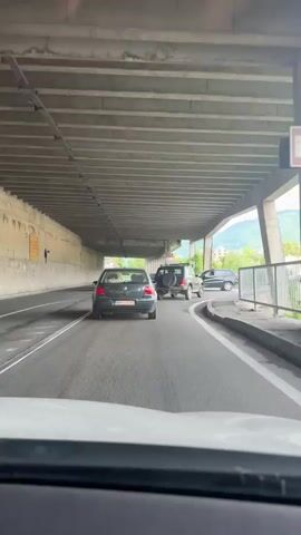 The video shows a car's perspective driving under a concrete overpass. There are other cars on the road ahead. The scene is a road with lane markings and a concrete barrier on the right. Trees and mountains are visible in the background beyond the overpass. Traffic signals and road signs are also present. The lighting suggests it is daytime.