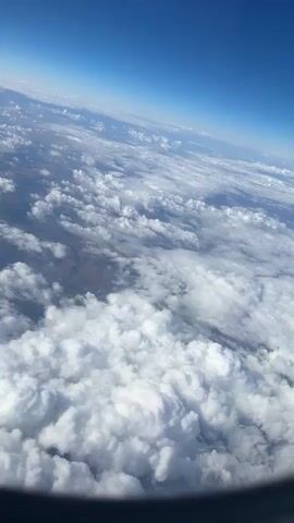 This is a static aerial photograph taken from an airplane window. The camera is angled downwards, looking out over a vast expanse of white, fluffy clouds. The clouds are dense and cover most of the lower half of the image, with some smaller, scattered clouds visible further in the distance. Below the clouds, the earth is visible as a muted landscape of browns and greens, with what appears to be a winding river or road cutting through it. The sky above is a clear, deep blue, gradually fading to a lighter shade towards the horizon. The bottom edge of the photograph shows the dark frame of the airplane window. There are no people or discernible objects other than the natural elements of sky, clouds, and land.