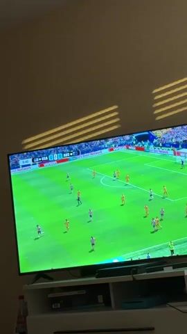 The media file shows a television screen displaying a soccer match. The game is between USA and AUS, with the score currently 0-0. The time displayed on the screen is 9:41 and 9:45, indicating the early stages of the match. The field is a vibrant green, with white lines marking the boundaries and penalty areas. Several players, dressed in white and red (presumably USA) and yellow (presumably AUS) jerseys, are scattered across the field. One player in the foreground, wearing a white jersey with red and blue stripes and the name "DEST" and the number "2" on the back, is facing away from the camera. The setting appears to be a living room, with a wall in the background and blinds casting striped shadows on the wall and the top of the television. Below the television, a white media console is visible, with some electronic devices and a bottle of Coca-Cola on the side. The lighting suggests it is daytime, with sunlight filtering through the blinds. There are no explicit cues about the weather, but the indoor setting makes it irrelevant to the visual. The overall mood is that of watching a sporting event, with no discernable emotions expressed in the static images. Visible text includes "Verizon" on advertising boards around the stadium, "USA 0", "AUS 0", and player names and numbers on jerseys.
