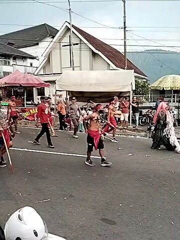 The video shows a street parade with several people dressed in traditional red loincloths and body paint, resembling a tribal or cultural performance. They are walking along a paved road, some carrying sticks or props. The background features buildings with a church spire, market stalls with tents, and flags, suggesting a public event or festival. Spectators are visible on the side of the road, some holding cameras or phones, indicating they are observing and documenting the procession. The sky is overcast. A motorcycle is partially visible in the foreground.