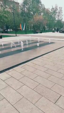 The video shows a public square on an overcast day. In the foreground is a water fountain with several small streams of water shooting upwards. The fountain is surrounded by light gray paving stones, with narrow drainage channels running along its edges. In the background, there are trees, benches, and a grassy area. Further back, a large, abstract, gray monument stands on a raised platform with steps leading up to it. A small bird walks near the edge of the fountain. A few people can be seen walking in the distance. Flags are visible in the background, near the trees.