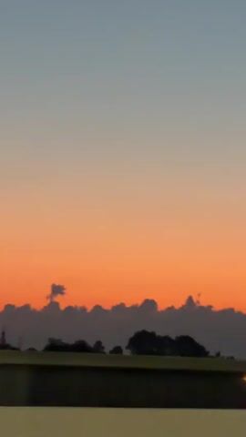 The video shows a moving shot of a sunset. The sky transitions from a pale blue at the top to a vibrant orange near the horizon. Silhouetted against the colorful sky are clouds and a dark, blurry line of trees and possibly some infrastructure along the ground. The camera appears to be in motion, possibly from a vehicle, causing a slight blur to the foreground elements. No people or distinct objects are clearly identifiable due to the distance and the low light conditions.
