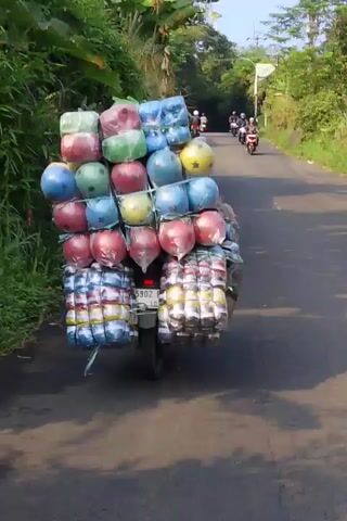 The video shows a motorcycle carrying a very large and precariously stacked load of goods. The goods appear to be colorful, round objects, possibly balls or balloons, wrapped in plastic. There are also bundles of what look like canned drinks or food items stacked at the bottom. The motorcycle is on a paved road with vegetation on either side. In the background, other motorcycles are visible, also on the road. The overall impression is one of a rural or semi-rural setting with a lot of cargo being transported by motorcycle. The load is extremely tall and wide, suggesting a significant amount of goods are being transported. The motorcycle has a license plate visible. The video appears to be taken from another motorcycle, as the perspective is slightly from the side and behind the main motorcycle.