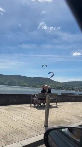 A wide shot shows a body of water with distant green hills under a blue sky with scattered clouds. Two kites are visible in the sky over the water. In the foreground, there is a paved walkway with a stone wall. Two people are sitting on a bench facing the water. To the left, a person walks by with a small dog on a leash. The camera is angled as if from inside a car, with a side mirror visible in the lower right corner.