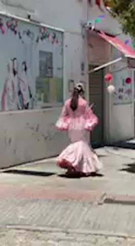 The video captures a bright, sunny outdoor scene, likely a street in Jerez de la Frontera, Spain, given the cultural context. The main subject is a person, possibly a flamenco dancer, dressed in a striking, voluminous pink gown. The dress features large, ruffled sleeves and a wide, flowing skirt that flares dramatically with movement.

The person is actively twirling and dancing, causing the expansive fabric of the dress to billow out, creating a dynamic visual effect. They move from left to right across the frame, then turn and dance back towards the left. The strong sunlight casts distinct shadows on the ground, highlighting the clear weather conditions.

In the background, there are white-washed buildings, consistent with Spanish architecture, one of which has a red awning and appears to be a storefront. Red and white decorative ornaments, possibly lanterns, hang outside this building. Towards the end of the clip, another person, partially visible in a brown jacket, walks past in the foreground. No discernible text is visible on any signs or objects. The scene conveys a sense of cultural celebration or artistic expression.