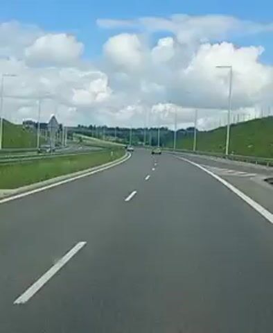 A first-person perspective video shows a view from the road of a highway with two lanes in the same direction. The sky is blue with scattered white clouds. A green hill is on the right side of the road. Streetlights are present on both sides of the road. There are a few cars visible on the highway. The road is paved and appears to be in good condition. The video is short and seems to be a snapshot of a journey.