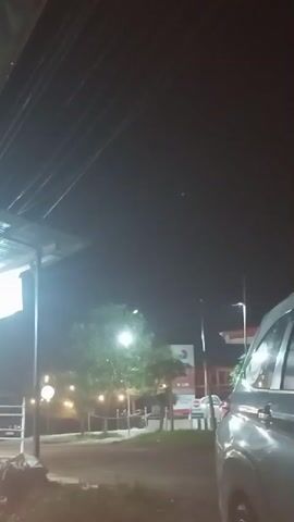 The scene is set at night, showing the side of a silver vehicle, possibly a van or SUV, parked near what appears to be a gas station or roadside service area. Wires are visible above, likely power or communication lines. A brightly lit tree is in the center, illuminating a small fenced-off area with decorative lighting. In the background, there's a gas station sign with a logo and additional road signage. The overall environment suggests a typical roadside stop during nighttime.