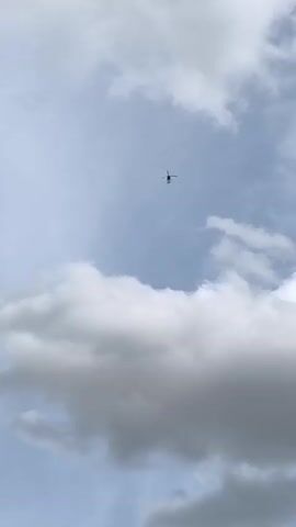 A black helicopter is seen flying in the sky against a backdrop of white and gray clouds. The helicopter is small in the frame and appears to be moving slowly from left to right. The lighting suggests it is daytime, possibly overcast.