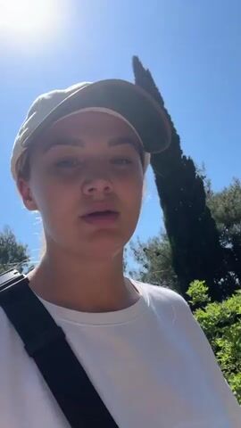 The video features a young woman on a hiking trail. She has blonde hair pulled back and is wearing a white long-sleeved shirt and a light-colored baseball cap with a darker brim. A black strap is visible across her chest, suggesting a bag or camera. The woman looks directly at the camera while talking, occasionally glancing at the trail ahead. The trail is rocky and surrounded by dense greenery, including bushes and trees, and the sky is bright blue. She briefly sticks out her tongue at the camera.