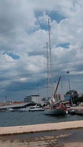 The video shows a harbor scene with a large sailboat prominently in the foreground. The sailboat is tall with multiple masts and rigging, and it has a red and white flag on its stern. Several other boats are visible in the background, including yachts and smaller vessels. The harbor appears to be busy with buildings and cranes along the waterfront. The sky is cloudy, with dramatic dark clouds suggesting an approaching storm or a recent downpour. The water is choppy, reflecting the overcast sky. The overall atmosphere is one of a working harbor with a mix of pleasure craft and commercial vessels, set against a moody sky.