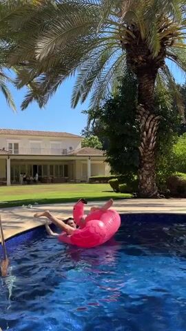 A person is floating on a pink inflatable flamingo in a swimming pool. The person is lying on their back, with their legs in the air. The pool is surrounded by a paved area and a grassy lawn. In the background, there is a large house with a palm tree in front of it. The sky is blue and clear.