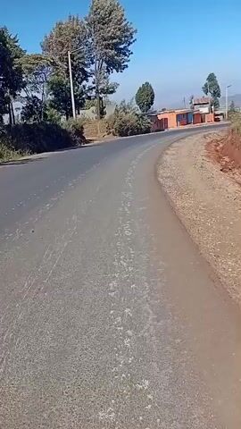 The video shows a paved road curving to the left. The road surface is a mix of asphalt and dirt, with some white markings visible on the asphalt. To the right of the road, there is a dirt shoulder with some gravel and a reddish-brown embankment. Beyond the embankment, there is a metal fence and some vegetation. In the background, there are buildings, trees, and a clear blue sky. The lighting suggests it is daytime.