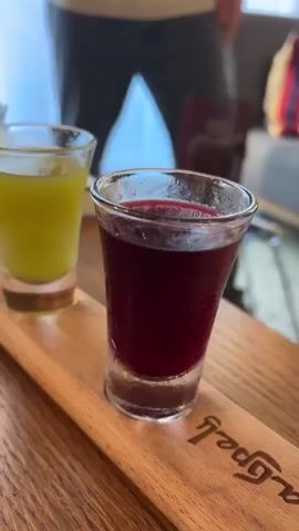 The video shows a wooden tray with four small glasses of colorful liquids. The glasses are arranged in a two-by-two formation. The liquid in the front left glass is a bright yellow. The liquid in the front right glass is a deep red. The liquid in the back left glass is a lighter yellow. The liquid in the back right glass is an orangey-red. The tray has text engraved on it, but it is difficult to read. The background is blurred but appears to be an indoor setting with natural light coming from a window.